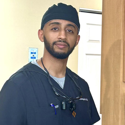 The image is of a man in scrubs, wearing a stethoscope and glasses, standing indoors with a serious expression.