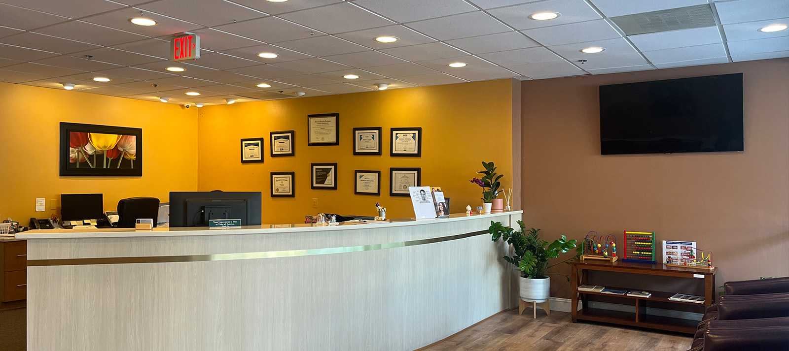 The image shows an interior view of a reception area, featuring a check-in desk with a computer monitor and keyboard, a framed diploma on the wall, and various decorative items.